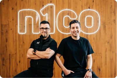 Dos hombres sentados frente a un letrero luminoso de n1co
